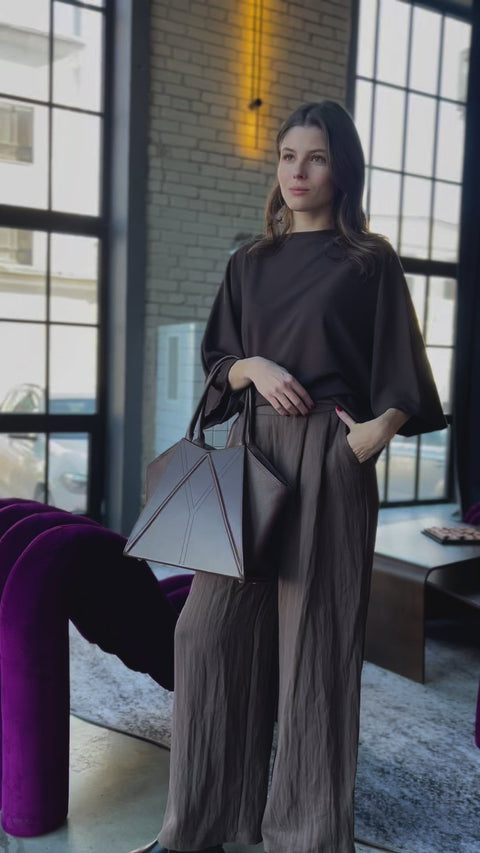 1. Woman holding Gerda Retter Design Leather Handbag Nova in caramel brown, styled with a green dress, against a concrete wall