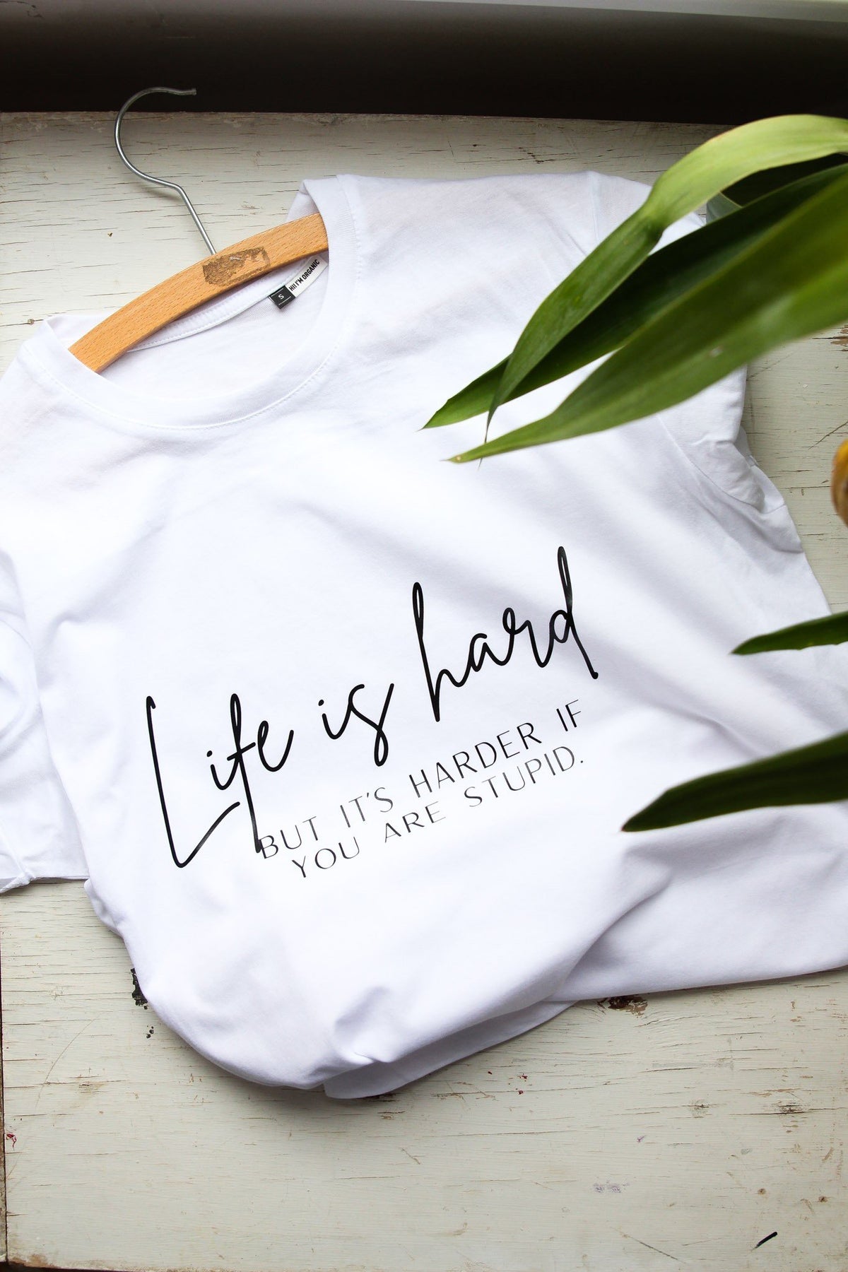 2. White organic cotton t-shirt with sarcastic text 'Life is hard but it's harder if you are stupid' on hanger, displayed on wooden background with plant detail