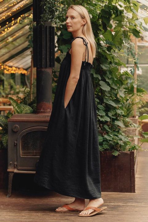 2. Side view of woman in black linen Ballade dress with inseam pockets in a lush indoor garden