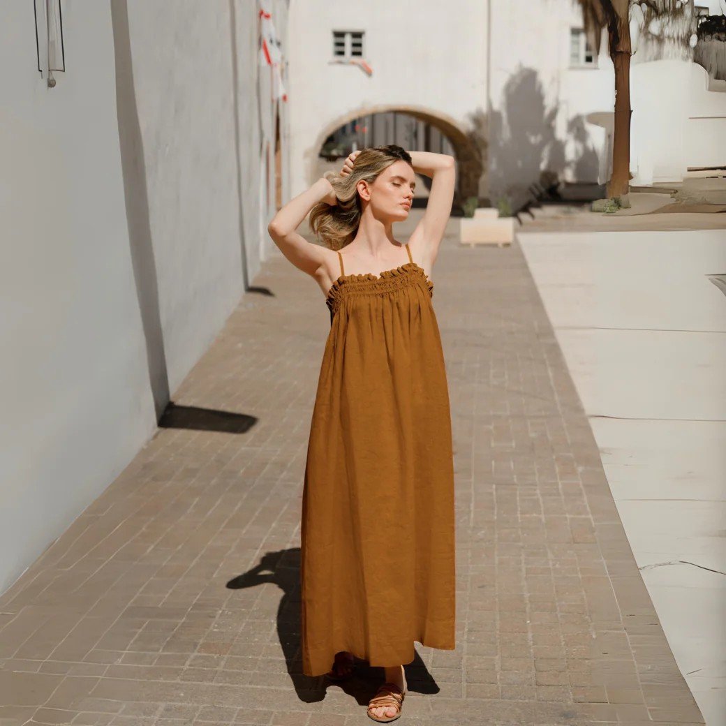 1. Woman wearing bronze linen Ballade dress with adjustable straps walking on a sunlit street, showcasing a relaxed summer style
