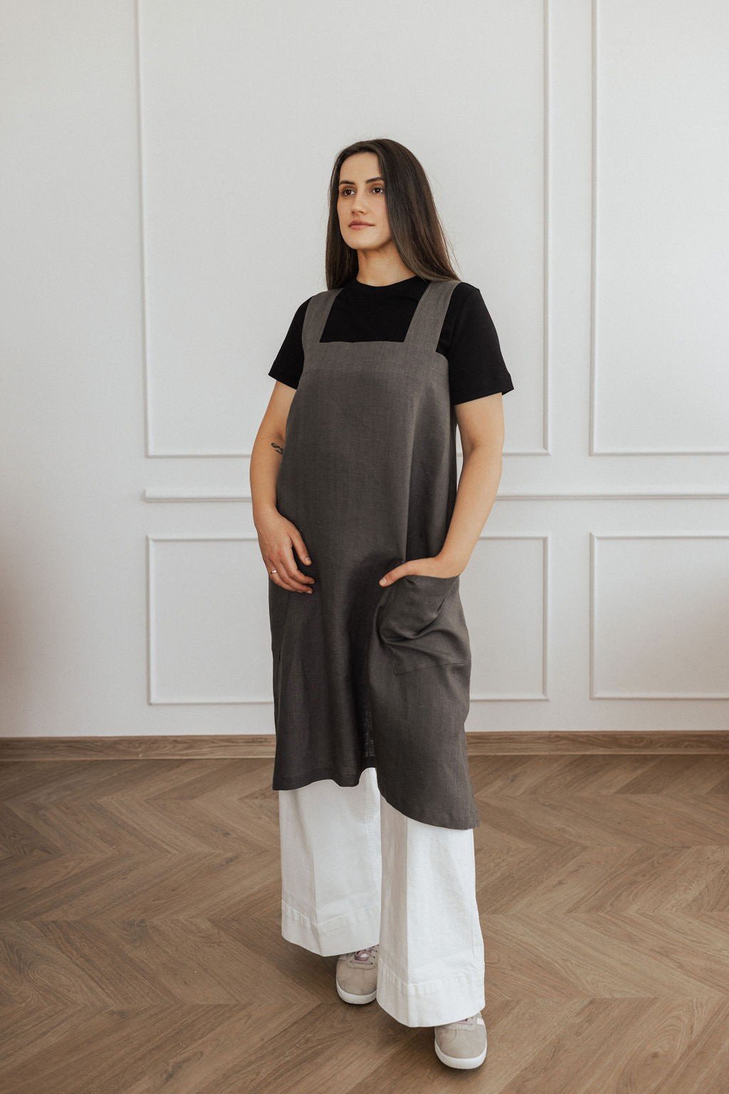 9. Woman in dark grey linen apron with hands in pockets, standing in a bright room