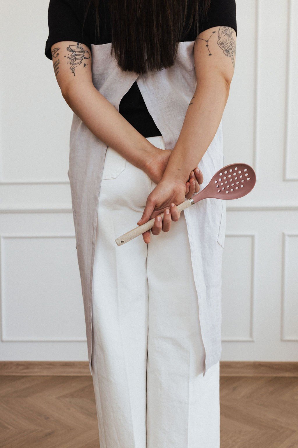 16. Back view of woman in light grey linen apron holding wooden spoon behind back