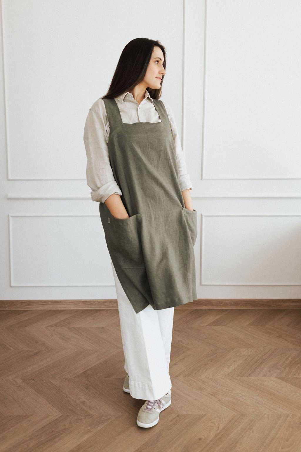 11. Woman in olive green linen apron with hands in pockets, standing in a bright room