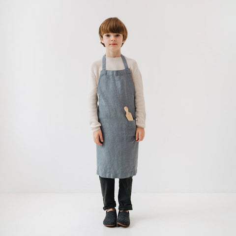 2. Child in blue fog linen apron with a wooden spoon in front pocket, standing against white background