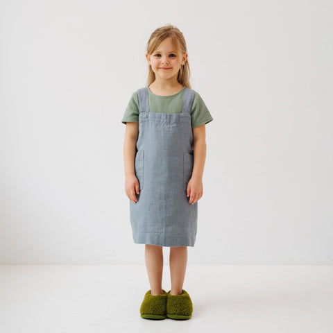 3. Girl in blue fog linen apron with green slippers, standing front-facing in a white room