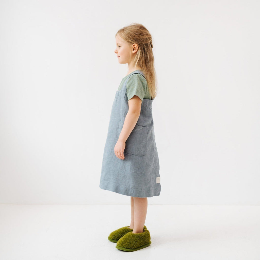 6. Side view of girl in blue fog linen apron with green slippers, standing in a white room