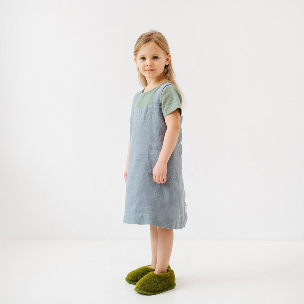 7. Girl in blue fog linen apron with green slippers, standing sideways in a white room