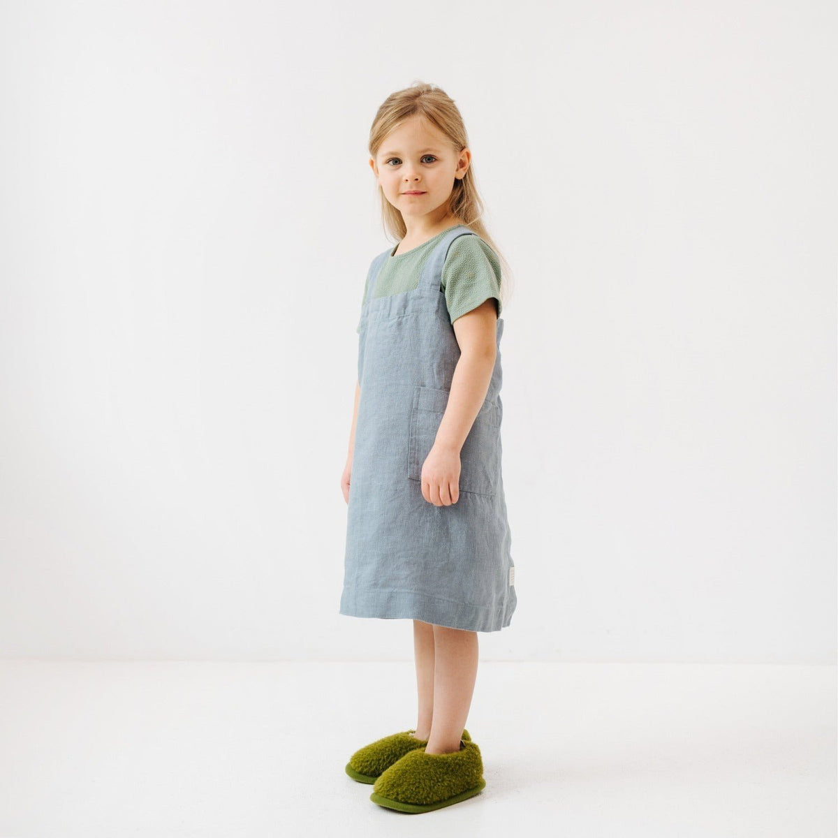 7. Girl in blue fog linen apron with green slippers, standing sideways in a white room