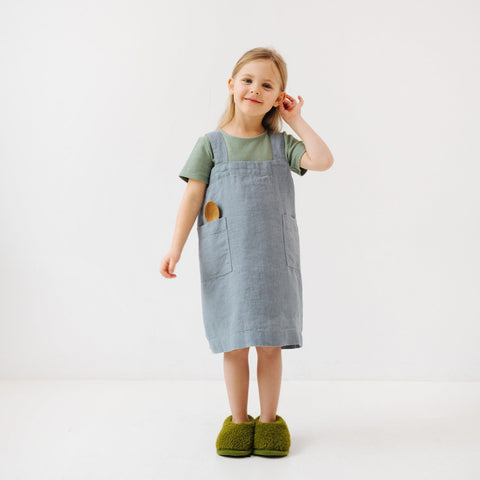 1. Girl wearing blue fog linen pinafore apron with green slippers, standing in a white room