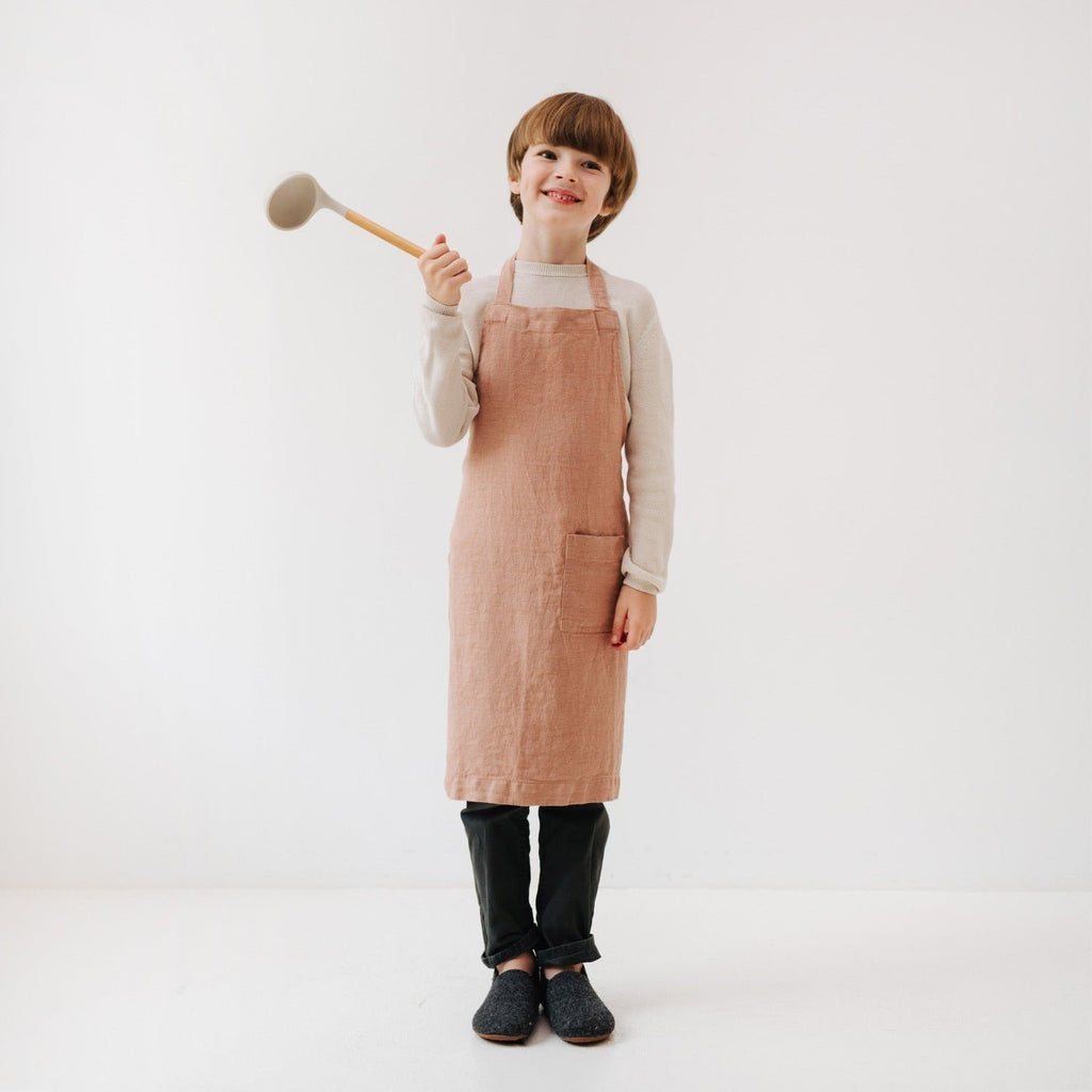 2. Child in cafe creme linen apron holding a spoon, smiling in a studio setting