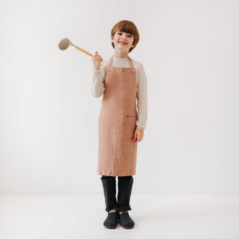 2. Child in cafe creme linen apron holding a spoon, smiling in a studio setting