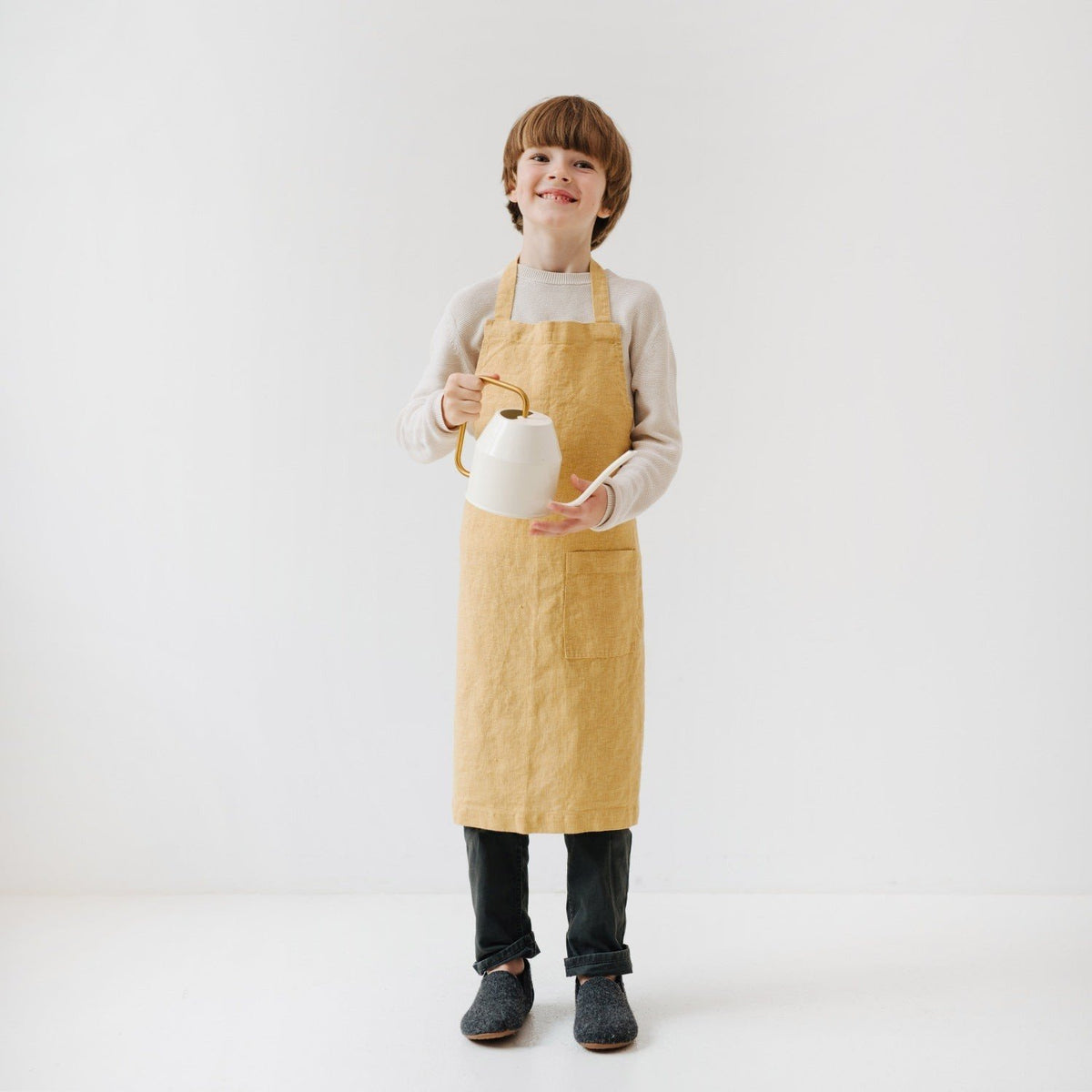 3. Child wearing honey-colored linen apron holding a white teapot in a bright, minimalist setting