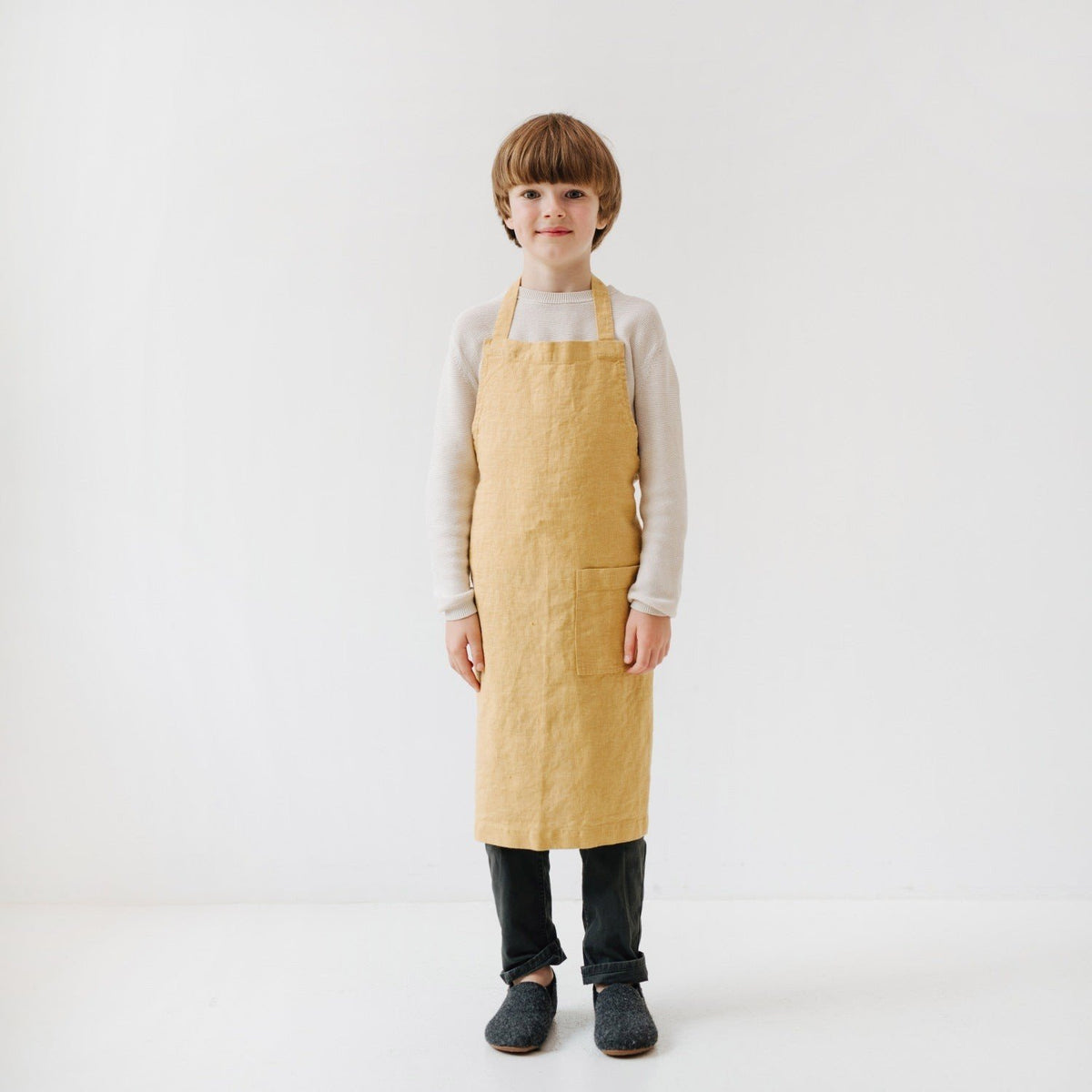 6. Child wearing honey-colored linen apron standing with hands by sides in a bright, minimalist setting