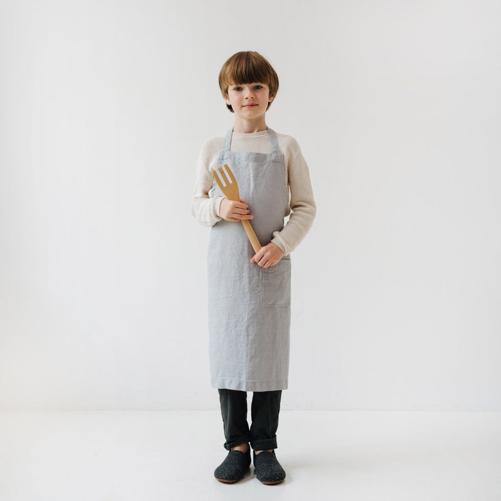 2. Child holding wooden kitchen tool while wearing light grey linen apron
