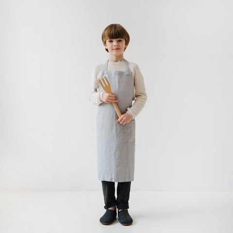 2. Child holding wooden kitchen tool while wearing light grey linen apron