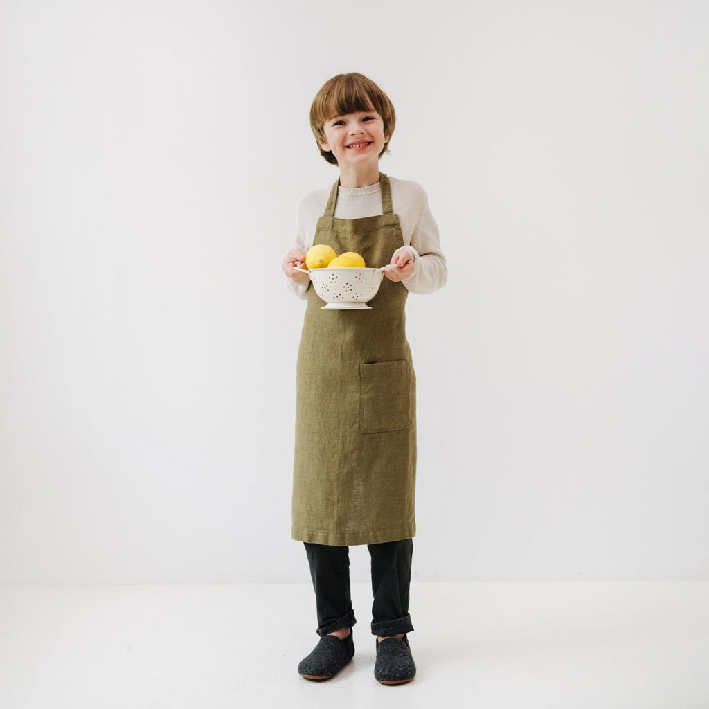 2. Child in olive green linen apron holding a colander with lemons, smiling in a white room
