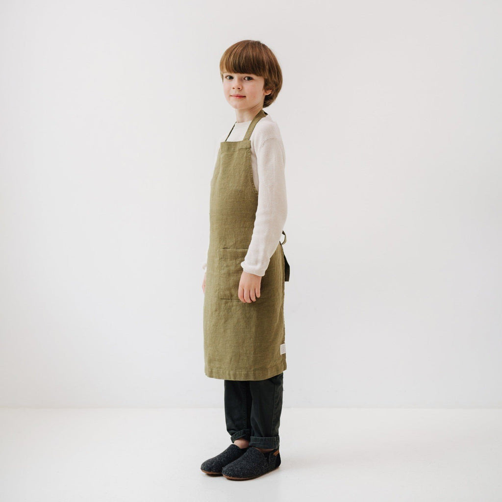 4. Side view of child wearing olive green linen apron with adjustable tie, standing in a white room