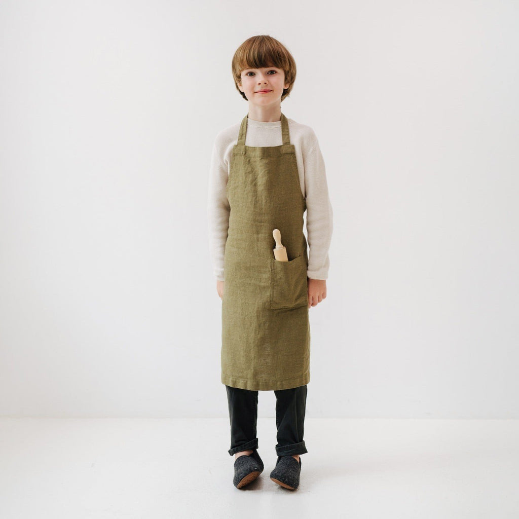 1. Child wearing olive green linen apron with front pocket, holding a wooden kitchen tool, standing in a white room