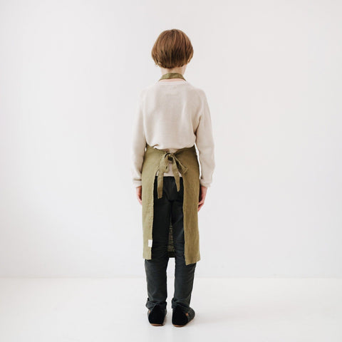 5. Back view of child wearing olive green linen apron with tied straps, standing in a white room