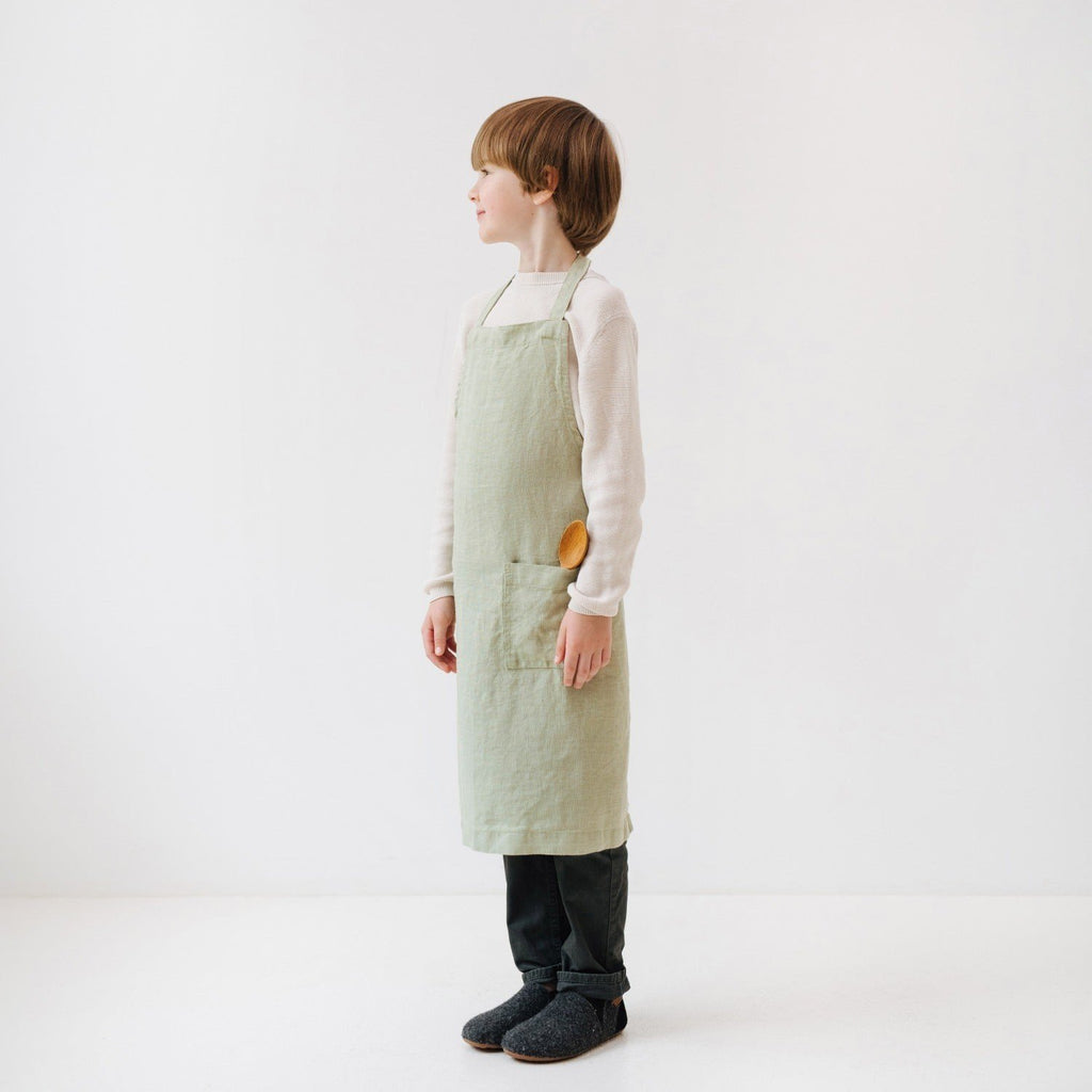 2. Side view of child in sage green linen apron with front pocket, standing in white studio