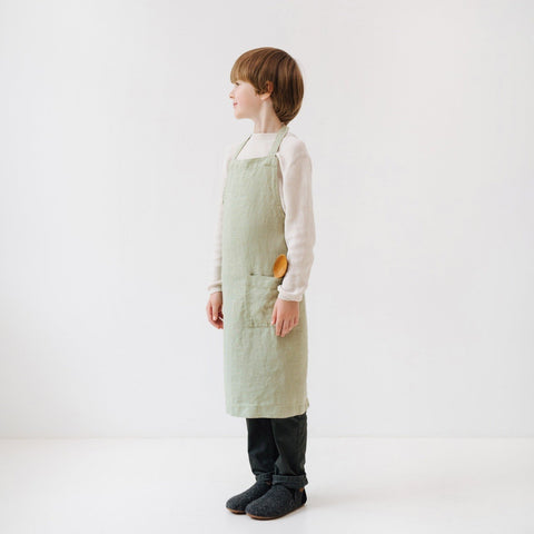 2. Side view of child in sage green linen apron with front pocket, standing in white studio