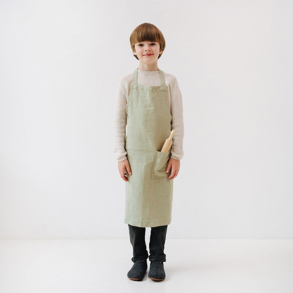 1. Child wearing sage green linen apron with front pocket, holding rolling pin, standing in white studio