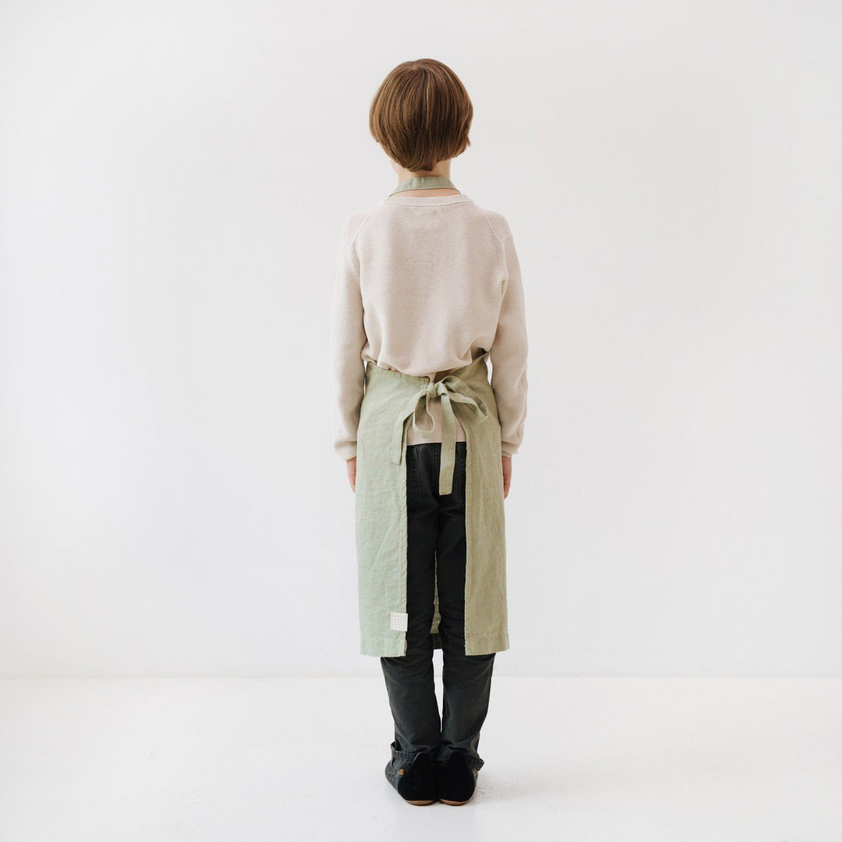 5. Back view of child wearing sage green linen apron with adjustable tie closure, standing in white studio
