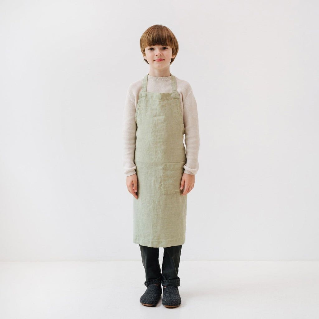 3. Front view of child wearing sage green linen apron with front pocket, standing in white studio