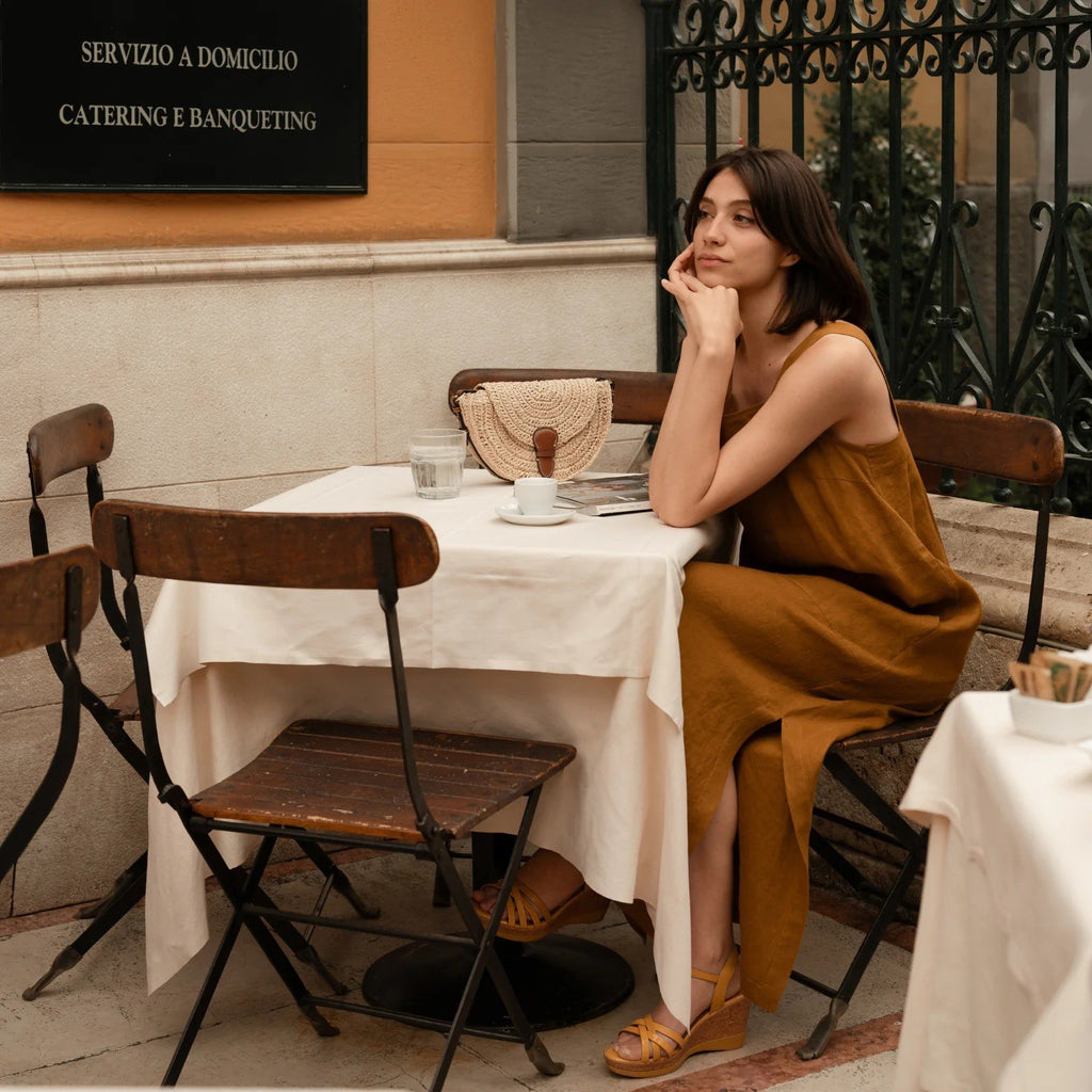 2. Woman in bronze linen dress sitting at an outdoor cafe table, resting her chin on her hand, with a woven clutch on the table