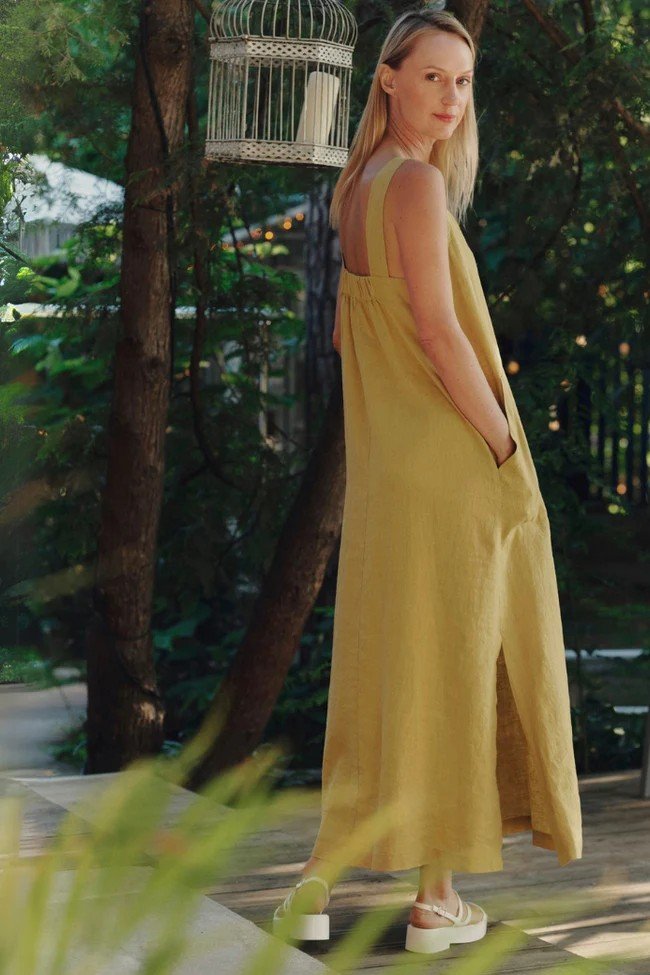 1. Woman wearing lemon curry linen dress with wide straps and side slits, standing outdoors near trees