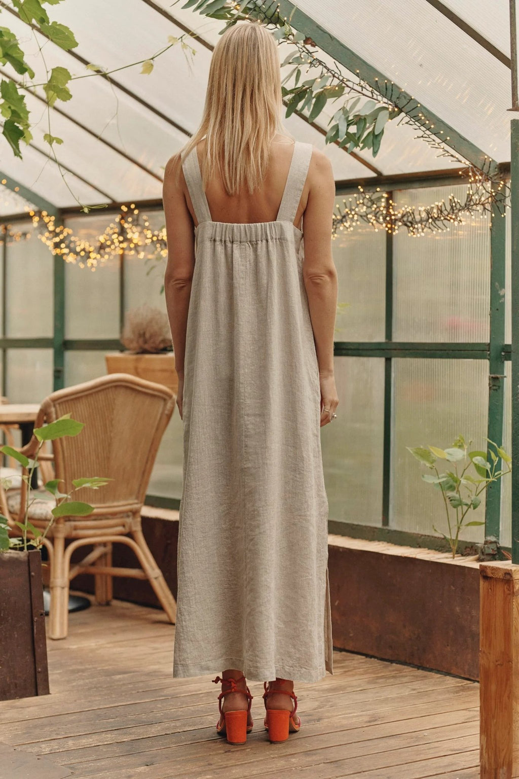 3. Back view of a woman in a long linen dress with elasticated back and wide straps, in a greenhouse setting
