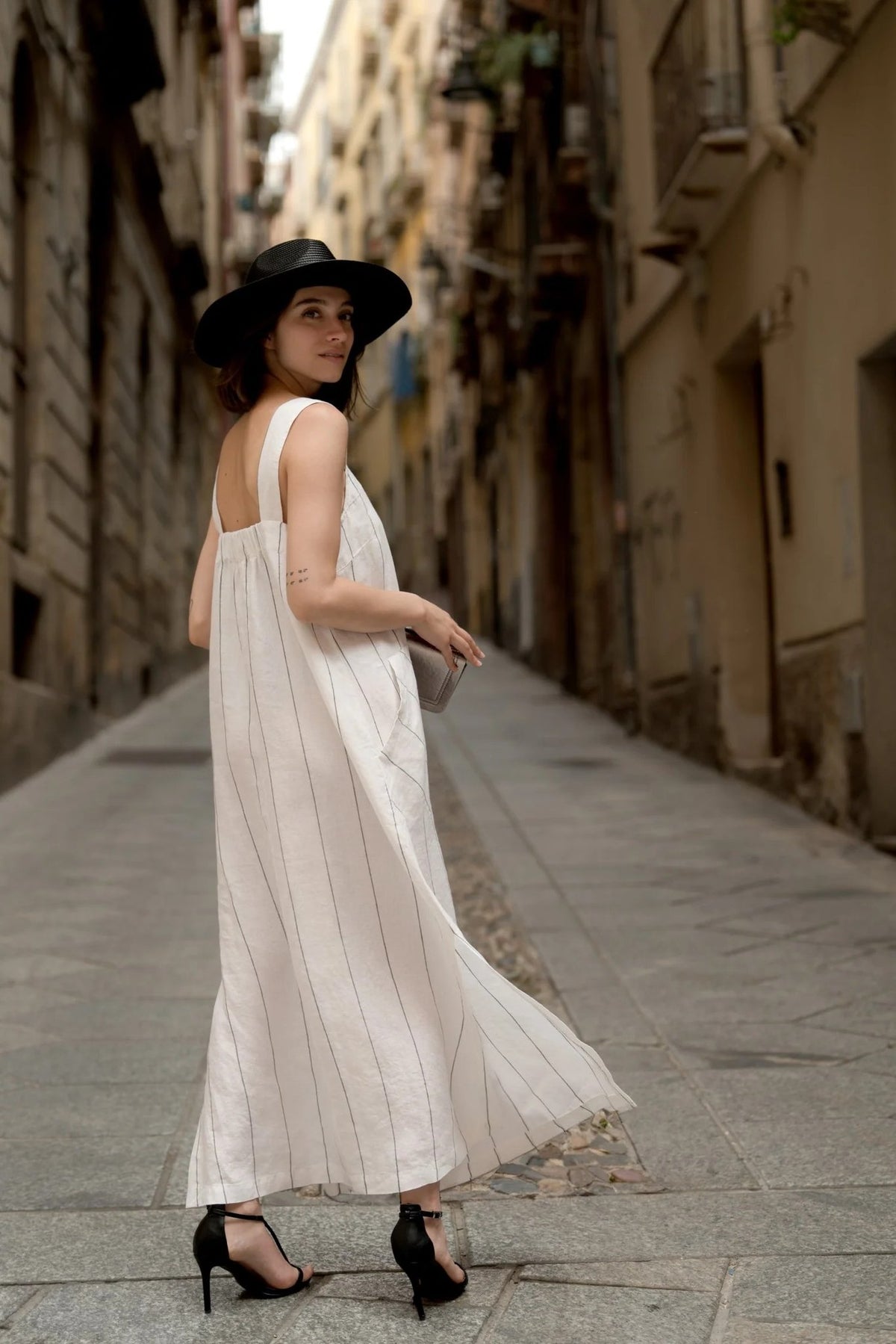 1. Woman wearing milk white linen dress with grey stripes and black hat walking down a narrow street