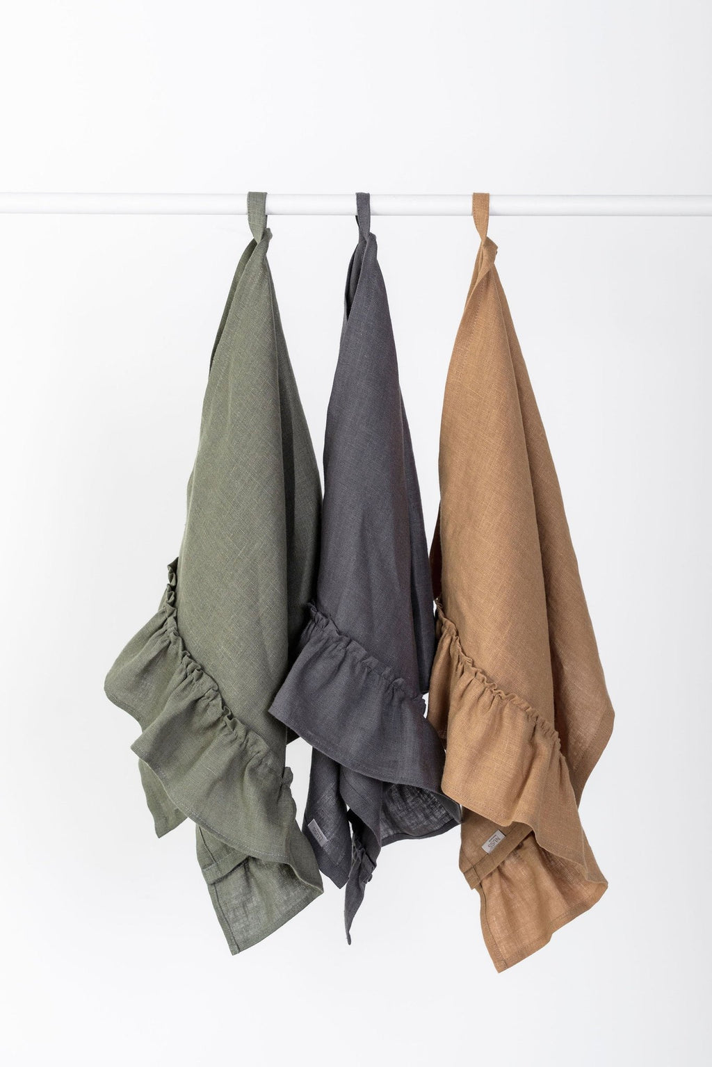 1. Three linen kitchen towels with ruffles in olive, gray, and caramel hanging on hooks against a white background
