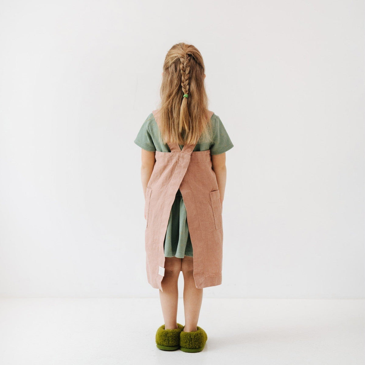 2. Back view of girl in pink linen pinafore apron with green shirt and braided hair in studio setting