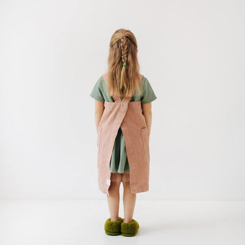 2. Back view of girl in pink linen pinafore apron with green shirt and braided hair in studio setting