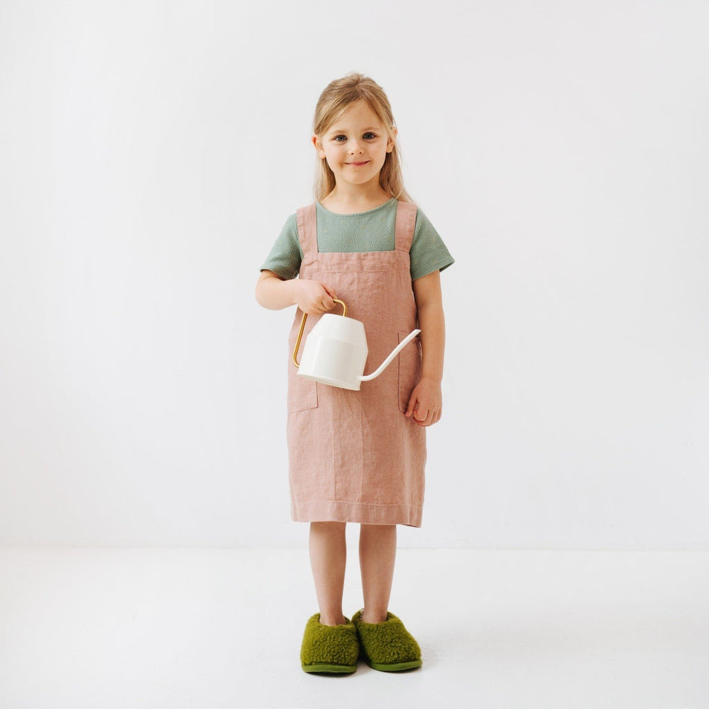 4. Girl wearing pink linen pinafore apron with green shirt holding white watering can in studio setting