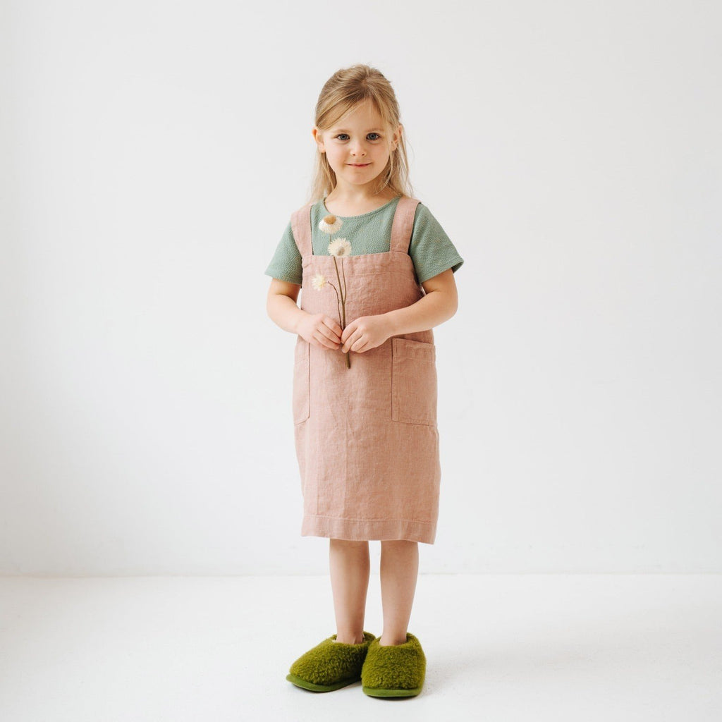 1. Girl wearing pink linen pinafore apron with green shirt and green slippers holding flowers in studio setting