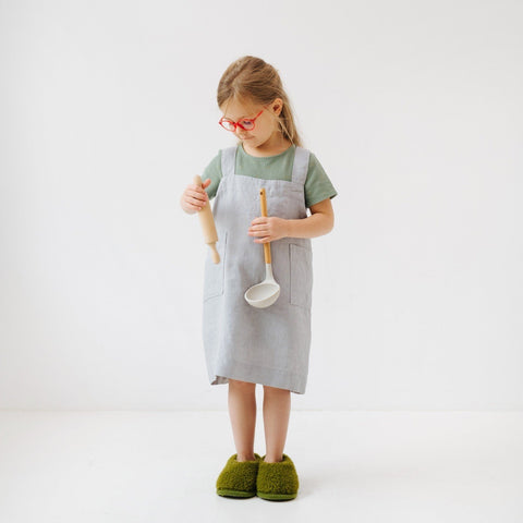 5. Girl in light grey linen pinafore apron holding a ladle and rolling pin, looking down