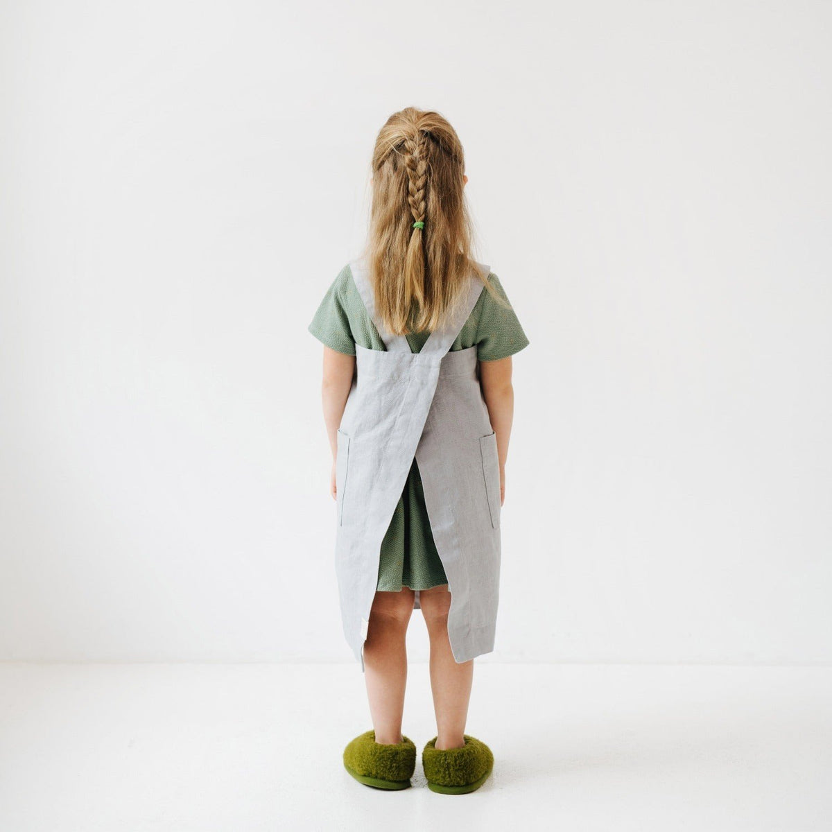 2. Back view of girl in light grey linen pinafore apron with green dress and slippers