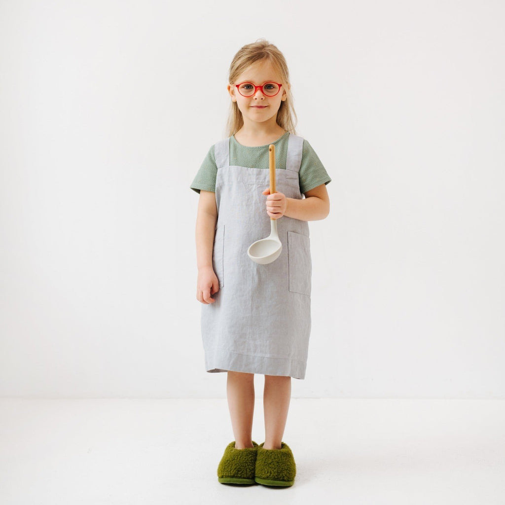1. Girl wearing light grey linen pinafore apron holding a ladle, standing in a white room
