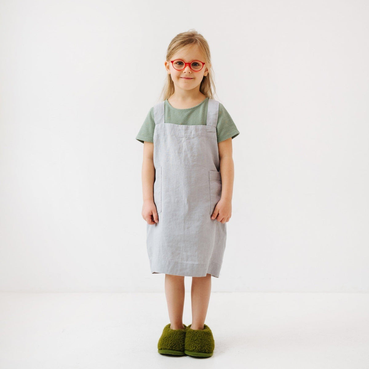4. Girl in light grey linen pinafore apron with green shirt and red glasses, standing in studio