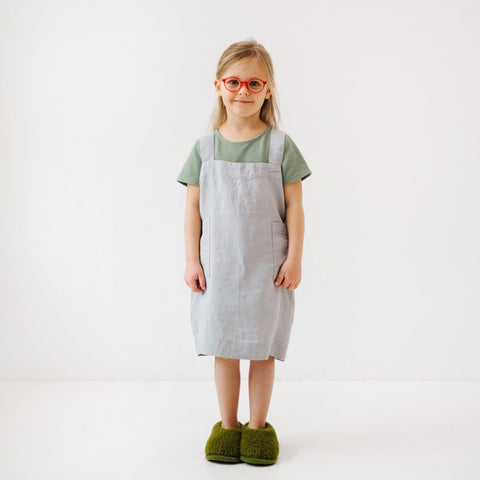 4. Girl in light grey linen pinafore apron with green shirt and red glasses, standing in studio