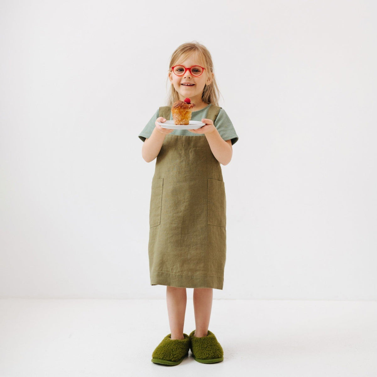 1. Girl wearing olive linen pinafore apron holding a plate with a muffin, standing on a white background