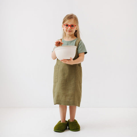 3. Girl in olive linen pinafore apron holding a mixing bowl, standing on a white background