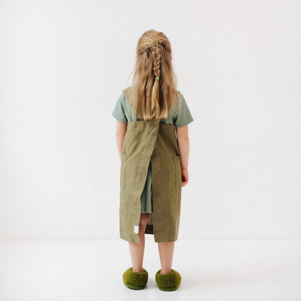 2. Back view of girl in olive linen pinafore apron with braided hair, standing on a white background