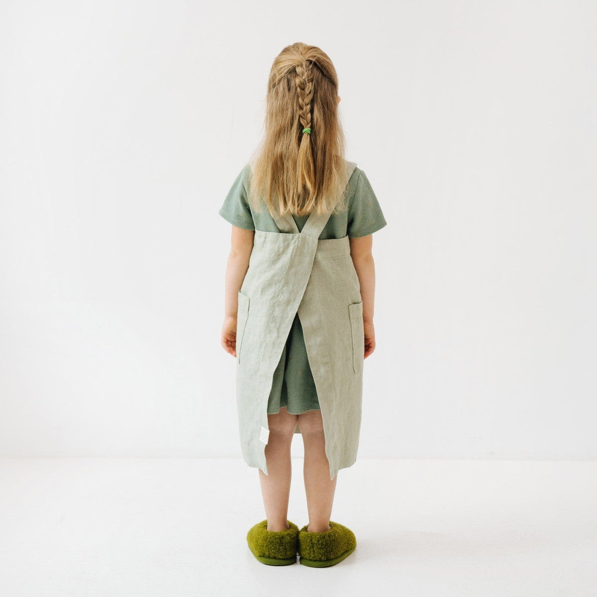 4. Back view of girl in sage green linen pinafore apron with braided hair, standing on white background