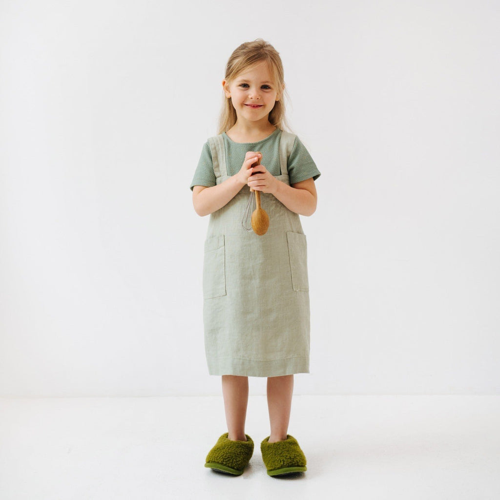 1. Girl wearing sage green linen pinafore apron holding a wooden spoon, standing on white background