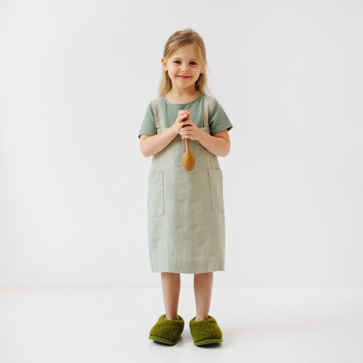 1. Girl wearing sage green linen pinafore apron holding a wooden spoon, standing on white background