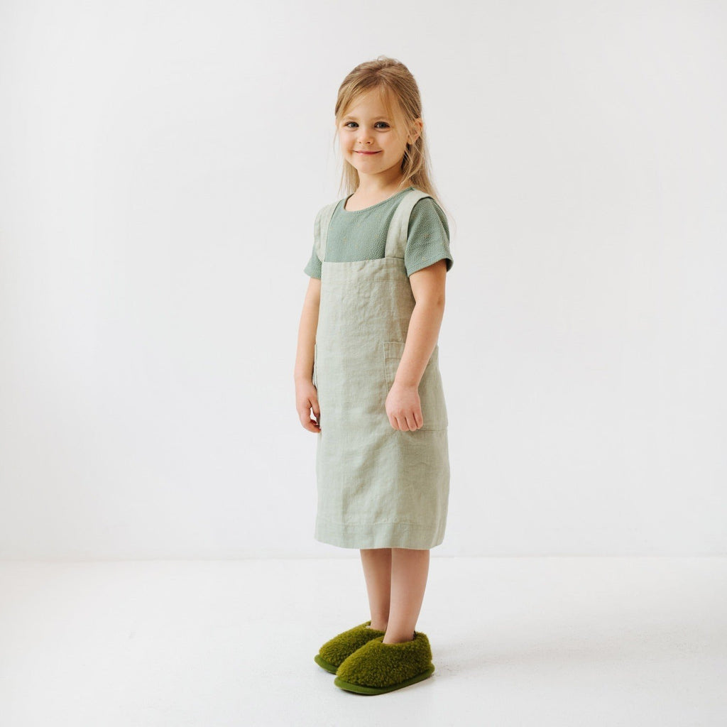5. Side view of girl in sage green linen pinafore apron smiling, standing on white background