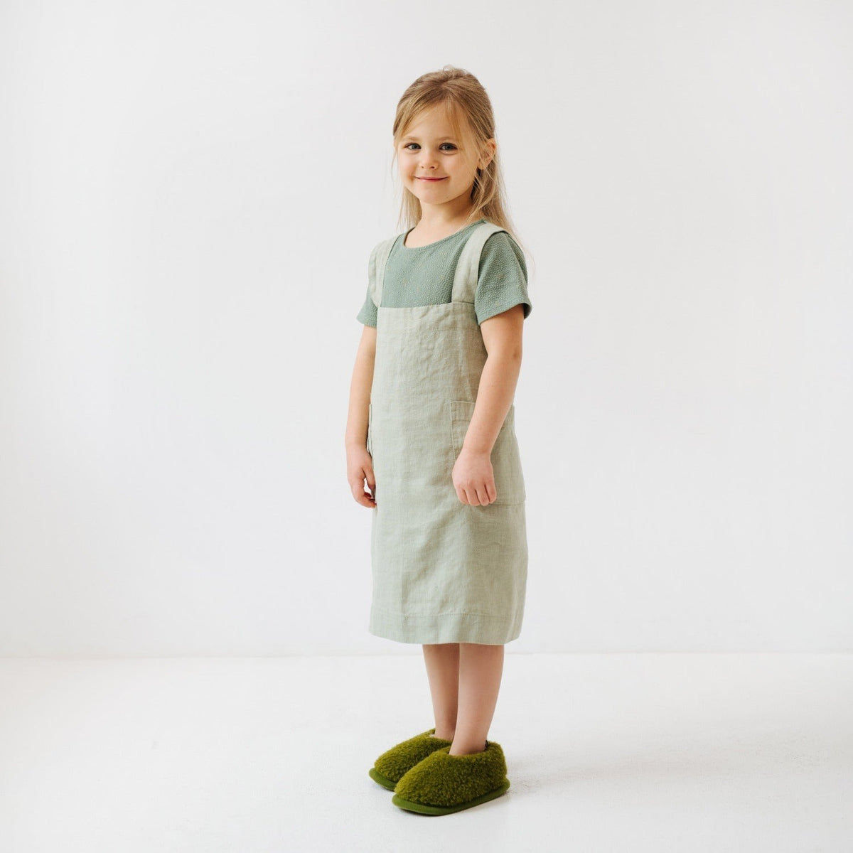 5. Side view of girl in sage green linen pinafore apron smiling, standing on white background